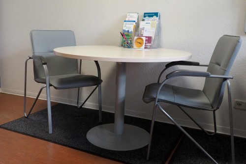 Tisch mit weißer Tischplatte und 2 grauen Stühlen. Einige Flyer sind auf dem Tisch ausgestellt.
